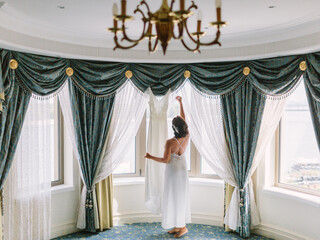 The bride in white stands at the bay window and holds her wedding dress. There are heavy green...
