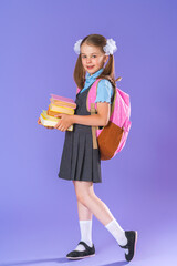 happy schoolgirl in uniform with backpack, walking along purple background in studio with a stack...