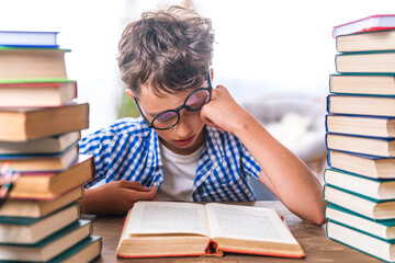 schoolboy tired of learning, wearing glasses, doing homework, sitting indoors between stacks of...
