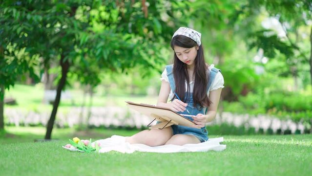 Portrait of attractive young pretty girl sitting in the public garden and drawing or painting a picture in summer season. Inspiration in green spring park. Art and relaxation on the holiday