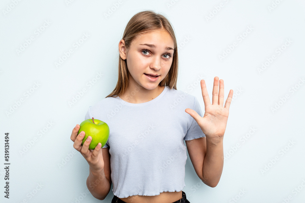 Wall mural young caucasian girl holding an apple isolated on blue background smiling cheerful showing number fi