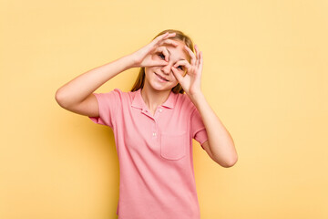 Young caucasian girl isolated on yellow background showing okay sign over eyes
