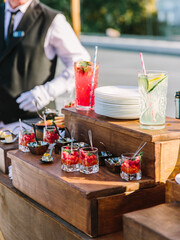 Obraz na płótnie Canvas Cocktails with strawberries, cucumber, and lemon, strawberry desserts in glass glasses stand on a wooden step rack on the street. In the background is a waiter.
