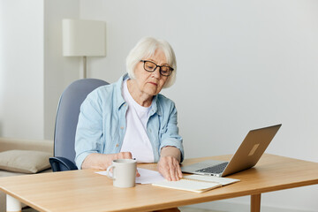 Fototapeta na wymiar a pleasant, stylish elderly woman is sitting at home in a bright interior and working on a laptop makes notes on a piece of paper. The concept of working from home