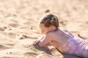 Portrait of a happy little girl, lying on the beach in summer on a sunny day. A child enjoys a summer children's holiday on the lake shore. Active summer holidays