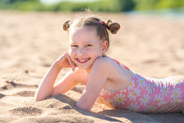 Portrait of a happy little girl, lying on the beach in summer on a sunny day. A child enjoys a summer children's holiday on the lake shore. Active summer holidays