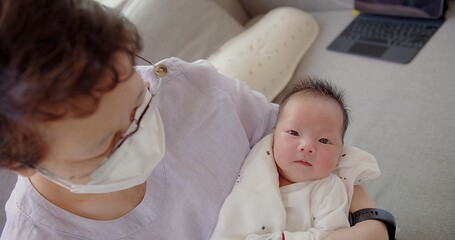 Happy Asian grandmother with protective mask talking looking at granddaughter newborn baby infant...