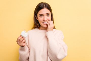 Young caucasian woman holding teeth whitener isolated on yellow background biting fingernails, nervous and very anxious.