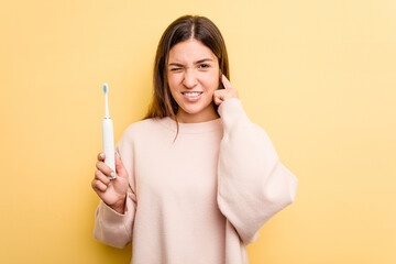 Young caucasian woman holding a electric toothbrush isolated on yellow background covering ears with hands.