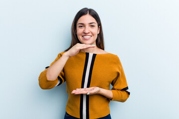 Young caucasian woman isolated on blue background holding something with both hands, product presentation.