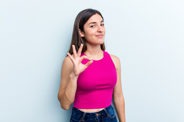 Fototapeta na wymiar Young caucasian woman isolated on blue background winks an eye and holds an okay gesture with hand.