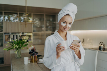 Happy woman in bathrobe and towel on head relaxing with cup of tea while doing cosmetic procedures at home