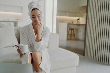 Happy woman in white bathrobe drinking tea and relaxing on sofa after taking shower or bath