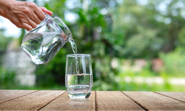 Water from jug pouring into glass on wooden table outdoors.Drink water pouring in to glass over sunlight and natural green background.Photo select focus with copy space.