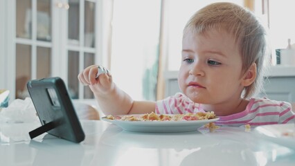 Cute Caucasian Toddler Baby Girl Eating Breakfast and Watching Video on Smartphone 
