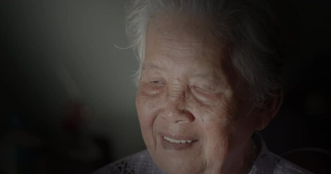 An Asian Woman Who Is A White-haired Elderly Person, Sits In A Dimly Lit House Talking On The Phone Alone With Her Distant Child With Loudspeaker On Because Her Ear Can't Hear Well.