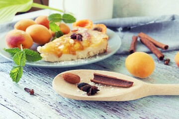 A piece of apricot pie on a plate, sprinkled with powdered sugar, spices and mint leaves, fresh apricots on the kitchen table, homemade food, sweet dessert