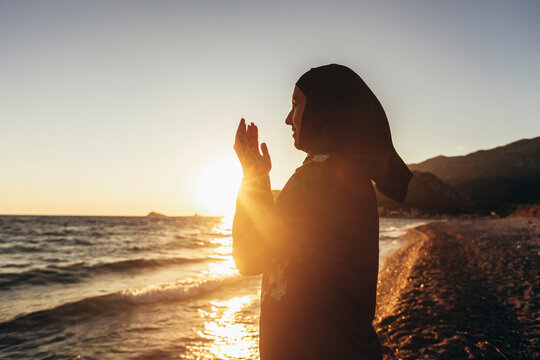 Muslim woman praying at sunset with hands up.