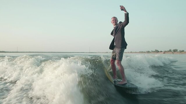 A surfer jumping on a wakeboard in a suit with a phone in his hands takes pictures of himself. A surfer takes a selfie and rides a wakeboard.