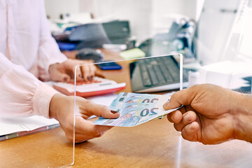 Hand of female social worker taking or giving euro money from client at office workplace with plexiglass separator. Counting grant subsidy rates. Government aid during financial crisis.