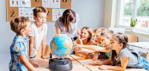 Happy cute enthusiastically pupils smiling around a globe in classroom with female teacher at...