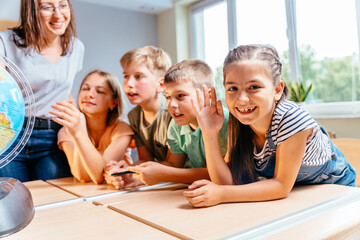 Cute enthusiastic little girl smiling, looking at camera and raising hand with teacher in class showing students a globe on background.