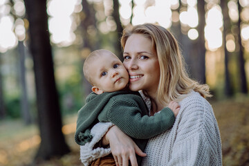 Mother holding her little baby son wearing knitted sweater during walk in nature, looking at camera.