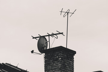 Black and white shot of an old antenna with dish next to brick chimney on the roof of an old house