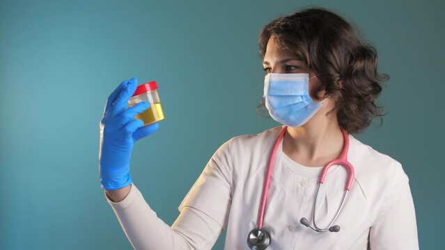 Woman doctor in a medical mask stands in a blue background and looks at urine tests.Jar of medical tests.Slow Motion