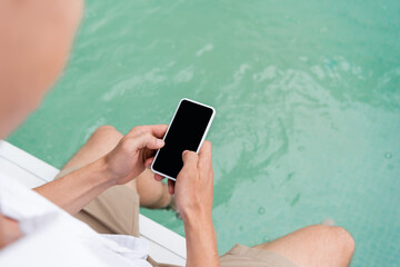 cropped view of man messaging on smartphone with blank screen near pool with turquoise water.
