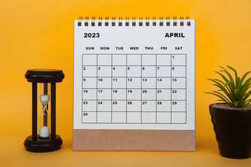 April 2023 desk calendar with table plant and minutes glass on yellow background.