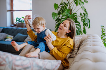 Young cheerful mother playing with her little child and having fun when lifting her up on sofa.