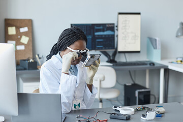 Portrait of young black woman wearing magnifying visor and inspecting hardware part in engineering...