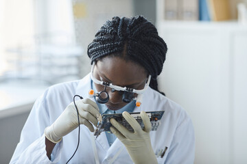 Portrait of young black woman inspecting computer parts and wearing magnifying visor while working in engineering lab