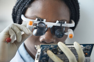 Close up of black female scientist wearing magnifying glasses and inspecting hardware part in...