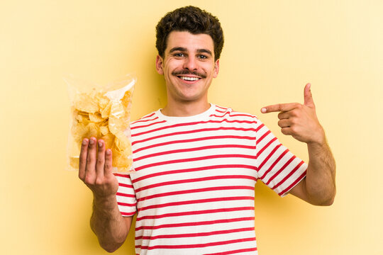 Young Caucasian Man Holding A Bag Of Chips Isolated On Yellow Background Person Pointing By Hand To A Shirt Copy Space, Proud And Confident