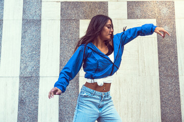 Photo shoot of stylish caucasian hip hop dancer posing having fun in blue jacket, jeans, black top and white tshirt, enjoy the urban town as background. City buildings shooting of moving fitness model