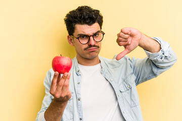 Young caucasian man holding an apple isolated on yellow background showing a dislike gesture,...