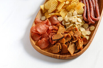 Wooden dish, plate with beer snacks, potatoes chips, meat chips, sausages kabanos, bread crackers. White background, copy space.