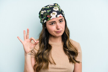 Young caucasian woman isolated on blue background winks an eye and holds an okay gesture with hand.