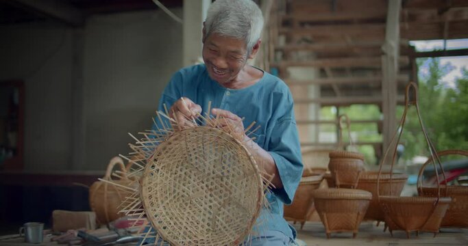 Slow motion scene of a happy smiling Asian male farmer who is an elderly native basketry artisan weave basket from bamboo that are cut into strips for weaving folk crafts in rural areas of Thailand.