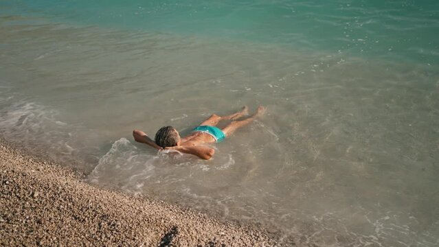 handsome athletic man in blue swim trunks relaxing by lying on pebble beach close to turquoise water of mediterranean sea in Oludeniz Beach in Aegean Sea after swimming and sunbathing. summertime wibe