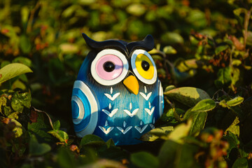 A toy of a surprised blue owl with multi-colored eyes close-up stands in the middle of green foliage in the light of the evening sun