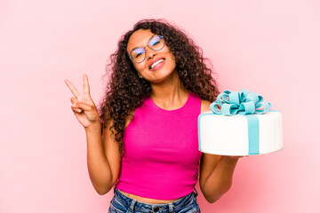 Young caucasian woman holding a cake isolated on pink background joyful and carefree showing a peace symbol with fingers.