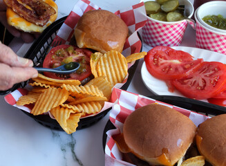 Adding toppings to Bacon Cheeseburgers with potato chips in a deli basket
