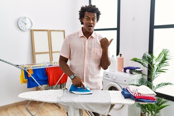 African man with curly hair ironing clothes at home surprised pointing with hand finger to the...