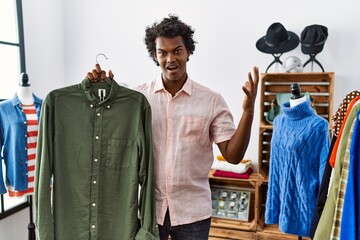 African man with curly hair holding shirt from clothing rack at retail shop celebrating victory with happy smile and winner expression with raised hands