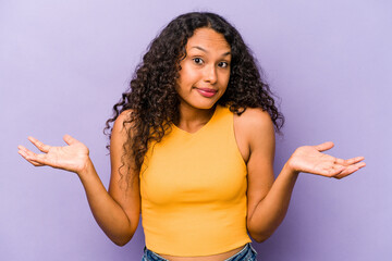 Young hispanic woman isolated on purple background doubting and shrugging shoulders in questioning...