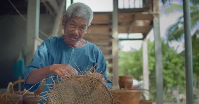 Slow motion scene of a happy smiling Asian male farmer who is an elderly native basketry artisan weave basket from bamboo that are cut into strips for weaving folk crafts in rural areas of Thailand.