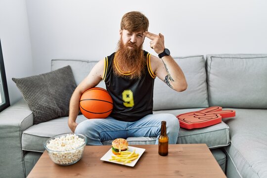 Caucasian man with long beard holding basketball ball cheering tv game shooting and killing oneself pointing hand and fingers to head like gun, suicide gesture.